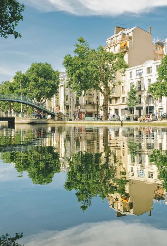 A scenic view of Canal Saint-Martin in the 10th arrondissement of Paris, with tree-lined walkways, a pedestrian bridge, and reflections of classic Parisian buildings in the water. A vibrant and trendy district popular among expatriates and real estate investors