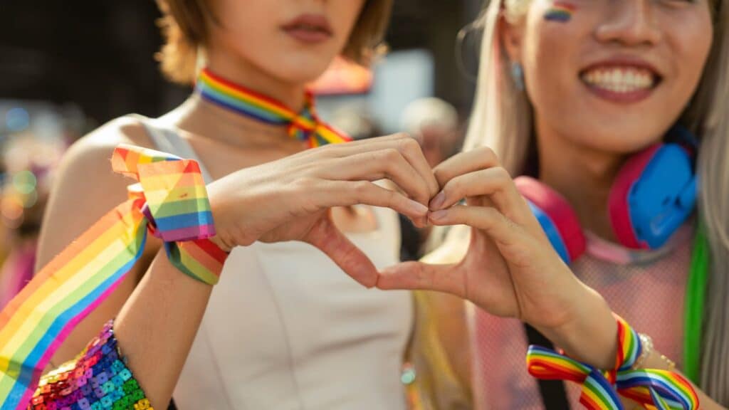 Two people making a heart gesture with their hands during a colorful event at Paris Gay Pride