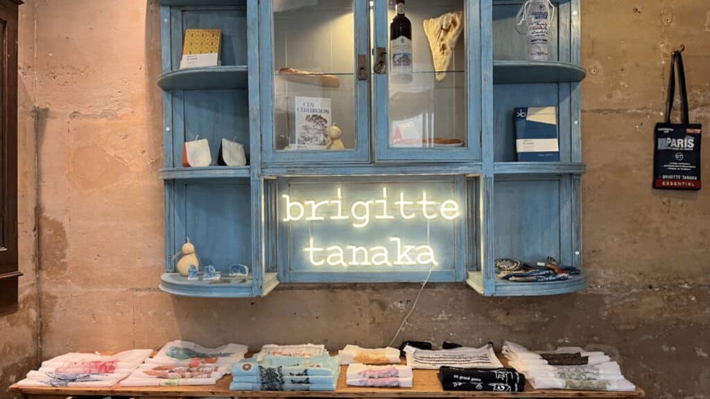 Rustic display with neon sign "Brigitte Tanaka" in Parisian boutique. Vintage blue shelves and assorted textiles below.