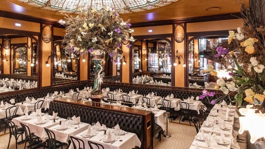 Ornate Parisian brasserie with decorative stained glass dome, elegant table settings, and floral centerpieces.