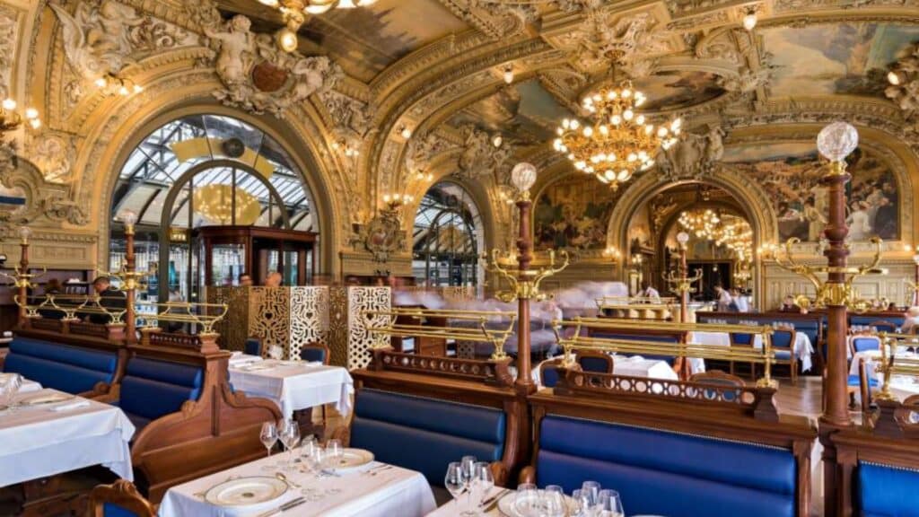 Ornate dining room of Le Train Bleu, Paris with chandeliers and mural ceilings. White tablecloths and blue velvet seating.