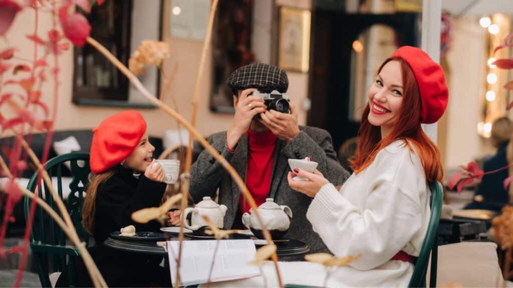 Family enjoying tea at Parisian café, wearing red berets; warm atmosphere with photographer capturing the moment.