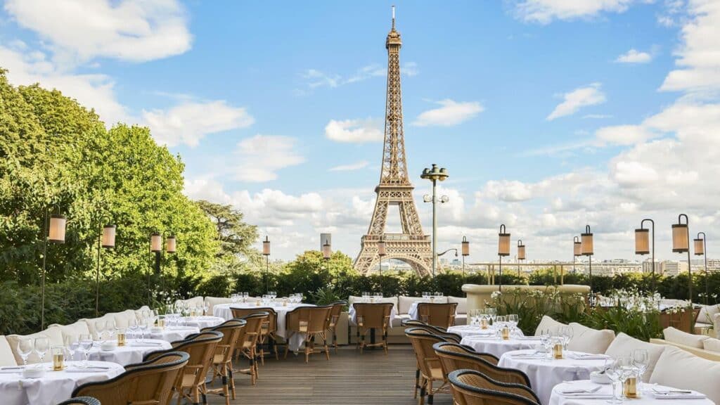 Outdoor Parisian cafe with Eiffel Tower view, elegantly set tables, wicker chairs, lush greenery, clear blue sky.