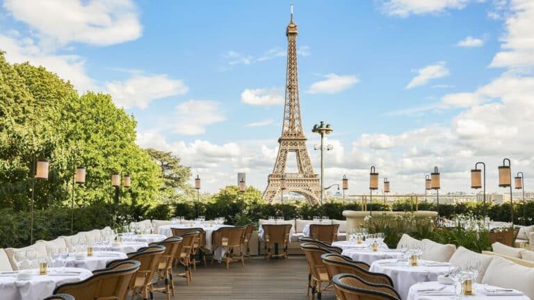 Outdoor Parisian cafe with Eiffel Tower view, elegantly set tables, wicker chairs, lush greenery, clear blue sky.