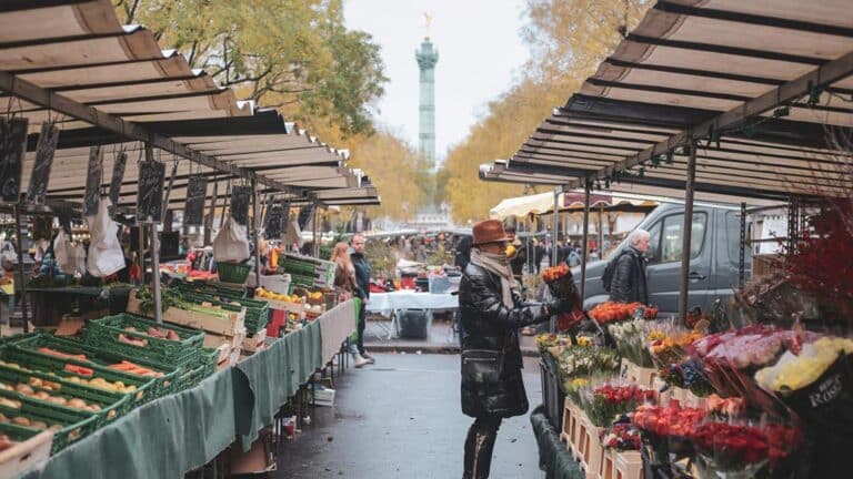 Outdoor Paris market with stalls, flowers, and produce; woman shopping; Colonne de Juillet in background.